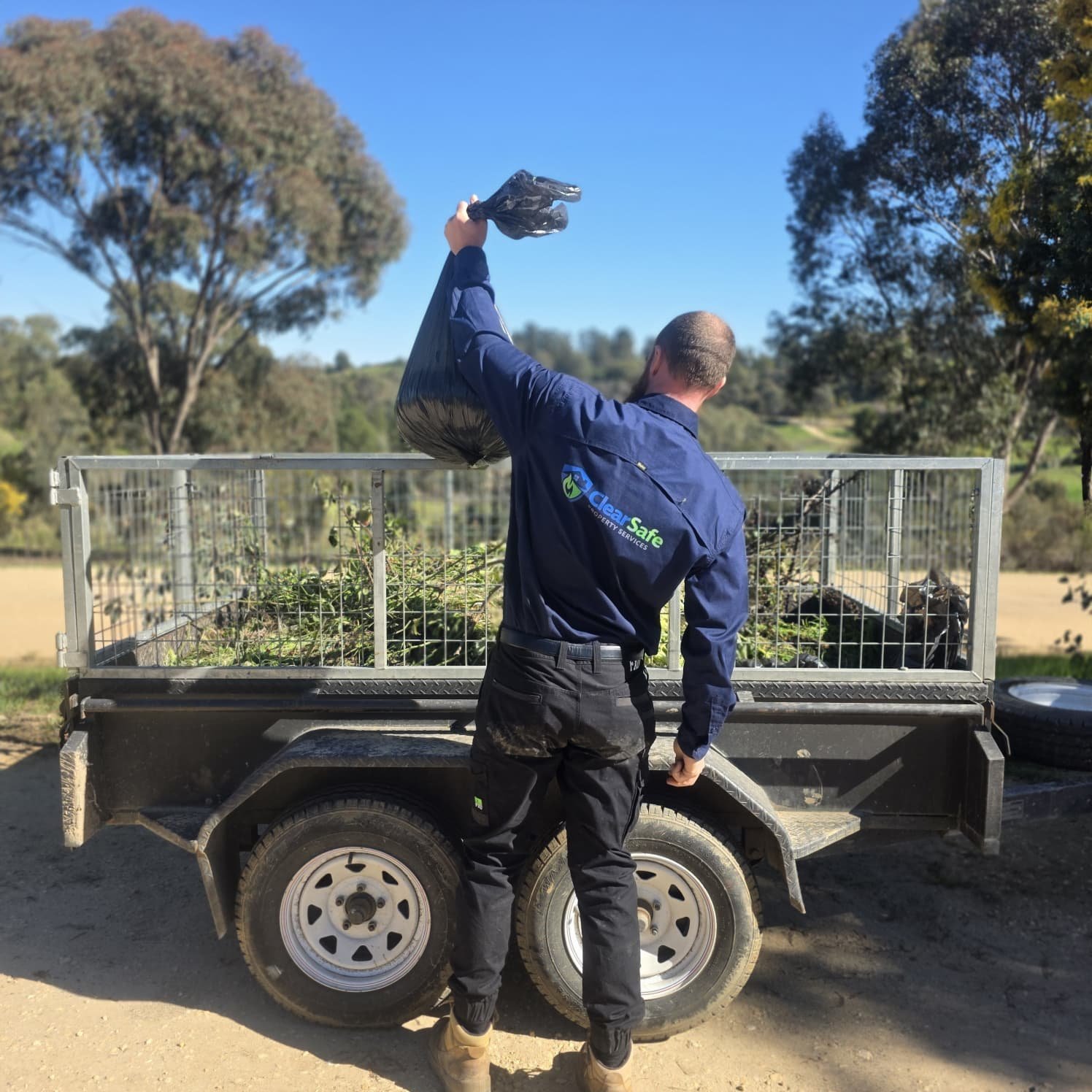 Worker in navy jacket standing on trailer holding equipment above head, rural landscape with trees in background